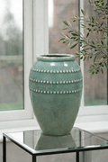 Green ceramic vase with decorative beads on a glass table in a room with large windows.