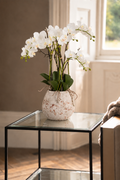 White artificial orchid flowers arranged in a distressed white ceramic vase with a twine bow, displayed on a glass-top side table in a softly lit room.