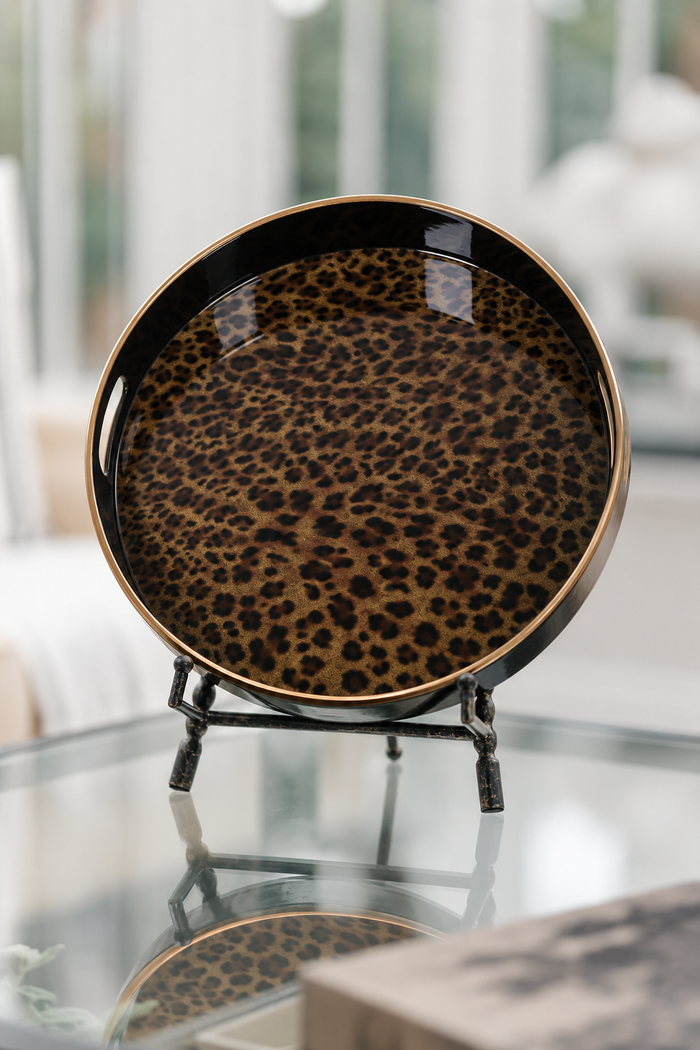 A round decorative tray with a leopard print pattern in brown and black, featuring a polished gold rim, displayed on a small black stand on a glass table.