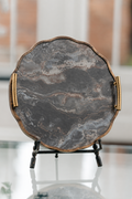 A scalloped oval serving tray with a dark gray and black marble pattern, featuring a bronze-toned metal rim and gold rope handles, displayed on a black stand.