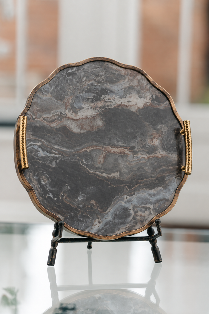 A scalloped oval serving tray with a dark gray and black marble pattern, featuring a bronze-toned metal rim and gold rope handles, displayed on a black stand.