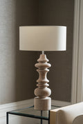A table lamp with a cylindrical off-white fabric shade and a sculptural, multi-tiered turned wood base in a light beige finish, shown on a glass-topped side table against a neutral gray wall.