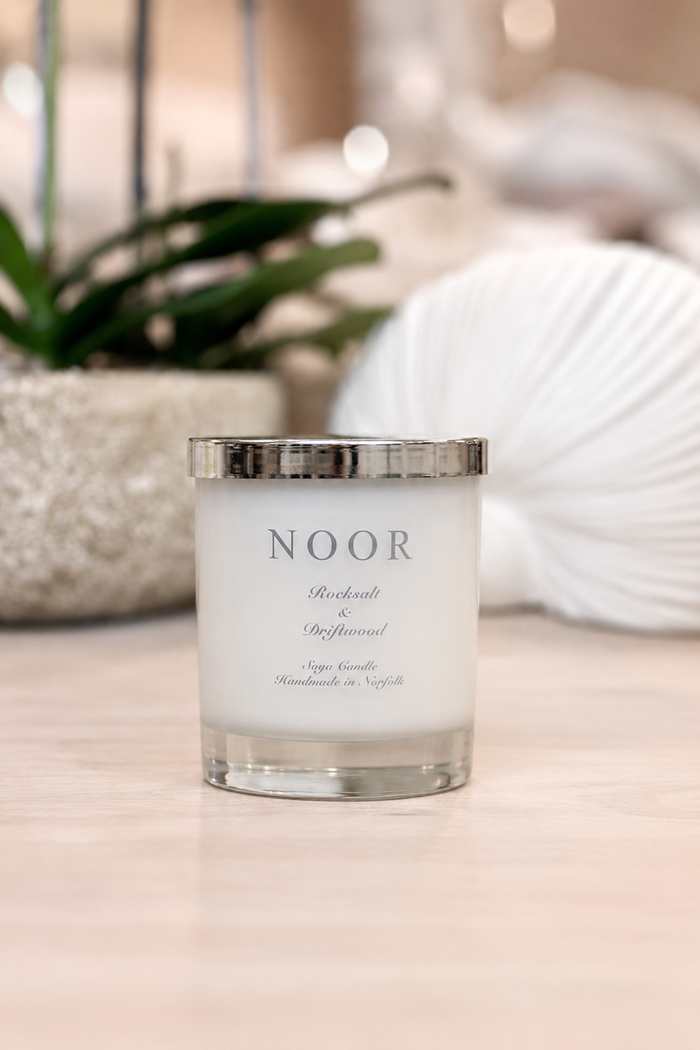 A white soy candle in a clear glass jar with a silver metallic lid, labeled 'NOOR', set on a light wooden surface with a blurred background of a plant and a white seashell.