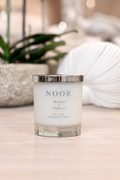 A white soy candle in a clear glass jar with a silver metallic lid, labeled 'NOOR', set on a light wooden surface with a blurred background of a plant and a white seashell.