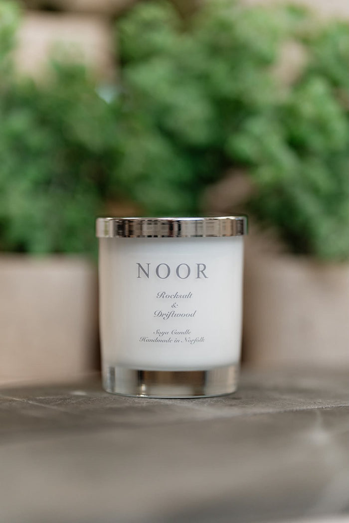 A white candle in a clear glass jar with a silver lid, labeled 'NOOR' and 'Absinthe & Spiced', set on a stone surface with a blurred green garden background.