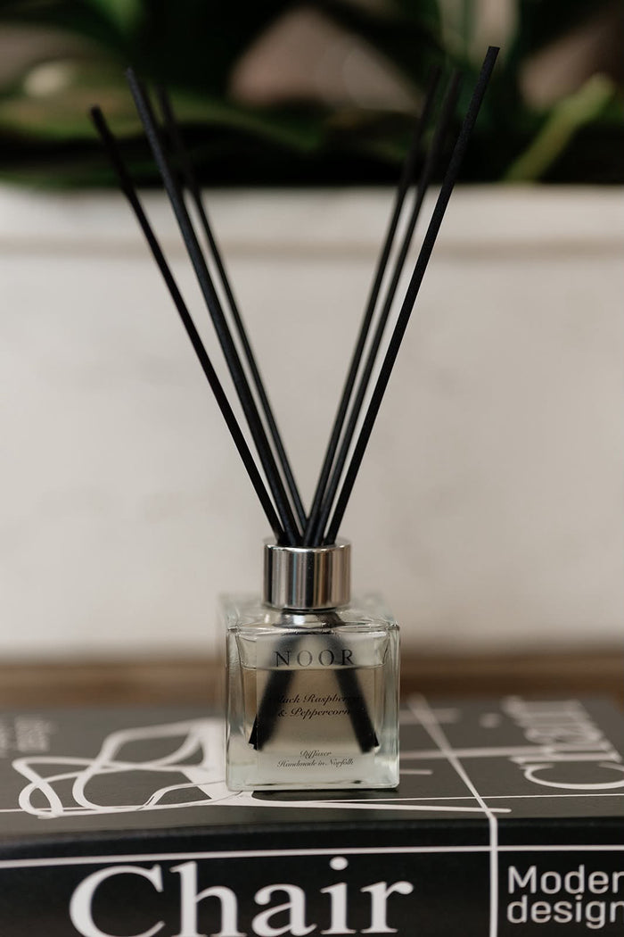 A clear glass reed diffuser bottle with black diffuser sticks, placed on top of a black book with the word 'Chair' visible, against a softly blurred indoor background.