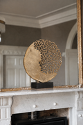 A round gold-toned decorative sculpture with an open, lace-like top half, displayed on a black rectangular base atop a white marble fireplace mantel in an elegant interior.