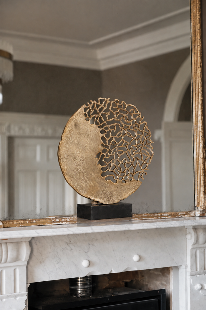 A round gold-toned decorative sculpture with an open, lace-like top half, displayed on a black rectangular base atop a white marble fireplace mantel in an elegant interior.