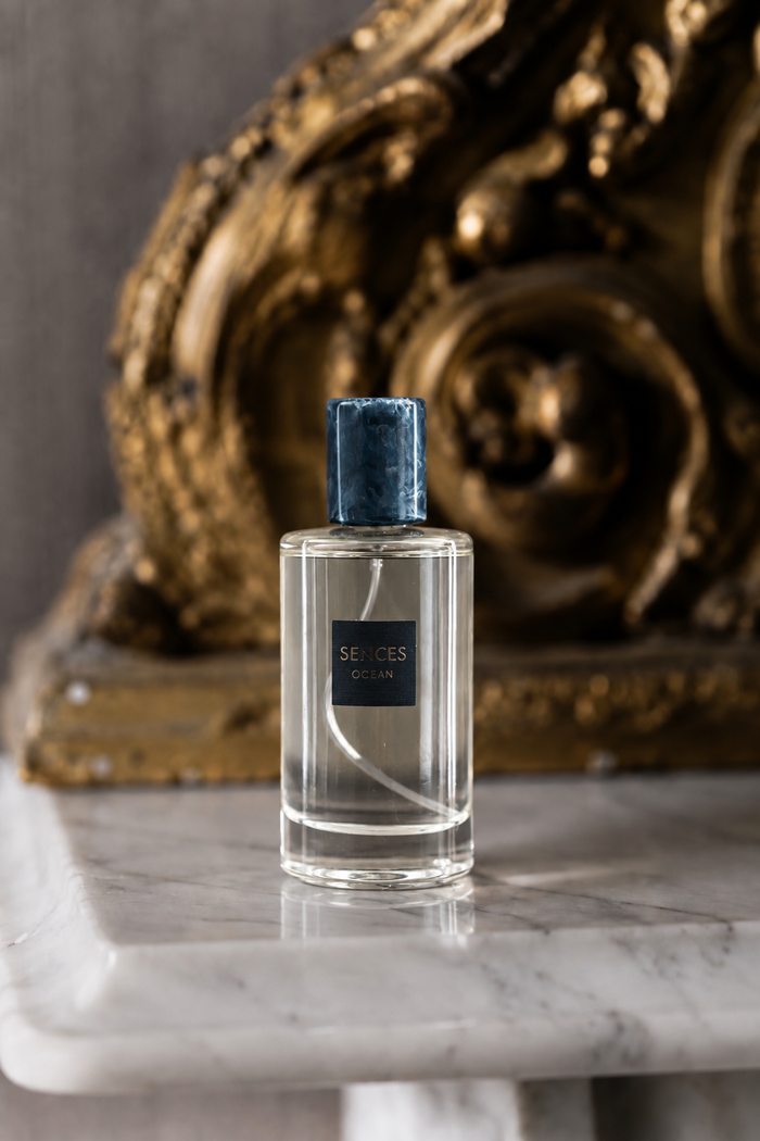 A clear glass perfume bottle with a dark blue velvet cap and a black label reading 'SEIGES CELINE', placed on a white marble surface in front of an ornate gold sculpture.