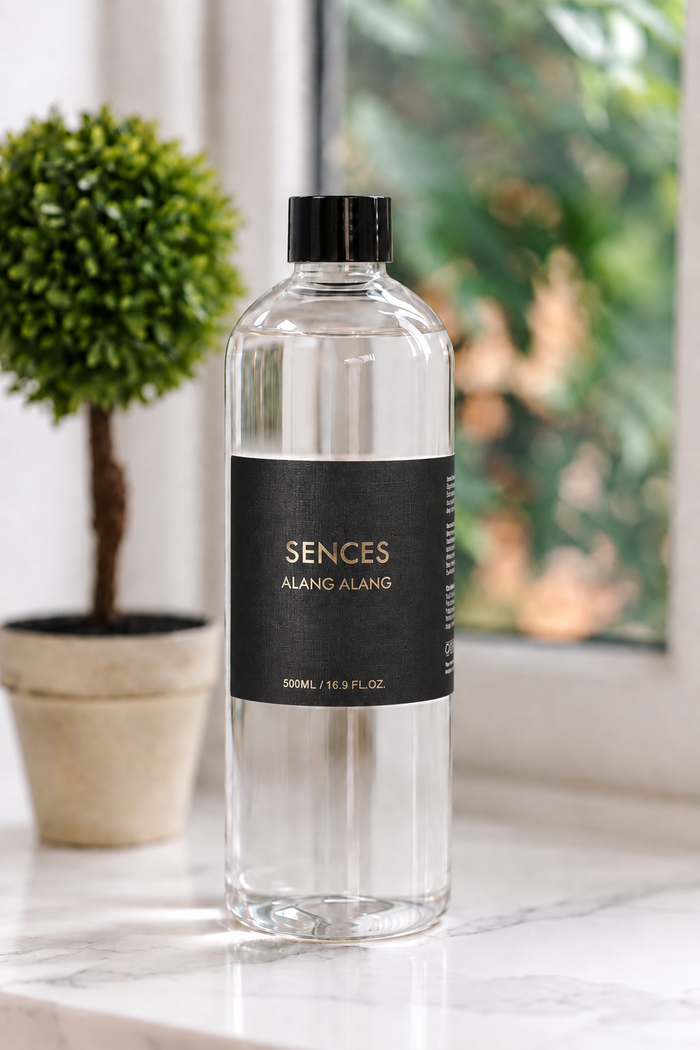A clear glass bottle of SENCES Alang Alang room spray with a black cap and black label, set on a marble countertop next to a potted plant in front of a window.