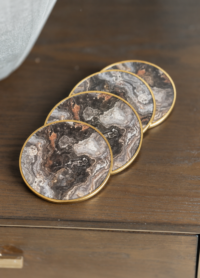 Four round coasters with a dark gray and black marble pattern and gold trim, arranged diagonally on a dark wooden surface.