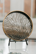A round decorative tray with a zebra-stripe pattern in black and metallic gold, displayed on a small black stand against a softly blurred indoor background.