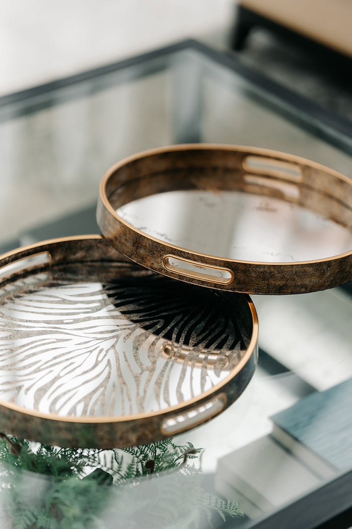 Two round decorative trays with distressed bronze metal rims and cutout handles, stacked on a glass tabletop with a blurred indoor background.